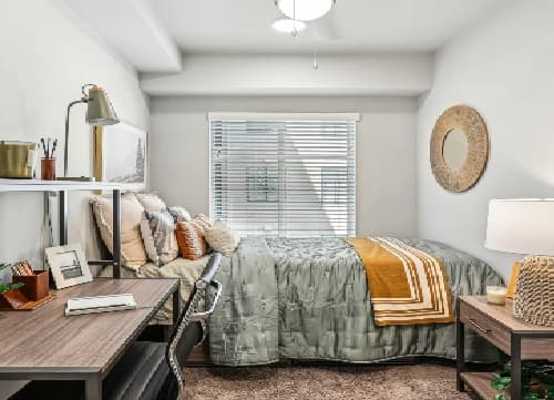A cozy bedroom at The Standard at Berkeley features a made bed with green and gold bedding, a desk and chair, lamp, and wall art. Sunlight streams through blinds, brightening the modern, tidy decor.