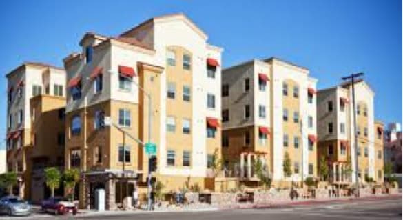 A modern, multi-story apartment building with tan and yellow walls, red awnings over the windows, and shops on the ground floor—an ideal option for SDSU housing set against a clear blue sky.