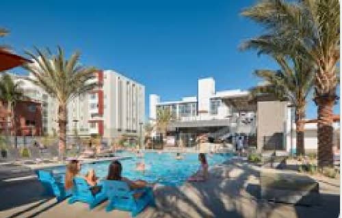 People relax in blue lounge chairs by a swimming pool surrounded by palm trees, with several modern SDSU housing apartment buildings and a clear blue sky in the background.