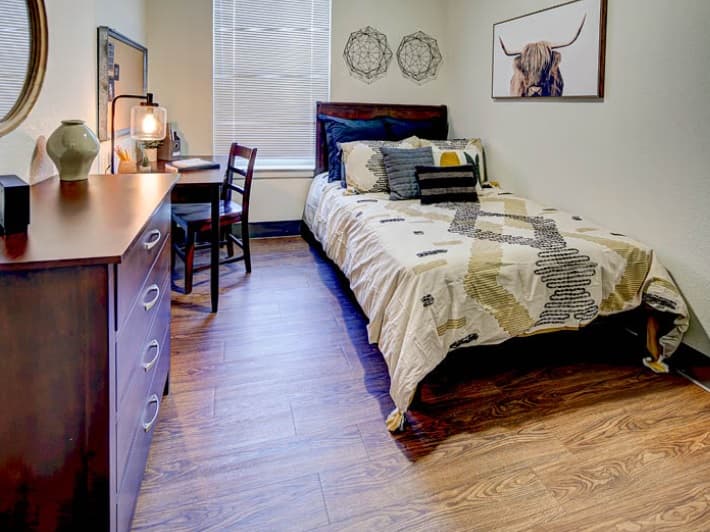 A tidy bedroom with a wooden floor, a bed with patterned bedding, a desk and chair by a window, a dresser with decor, a lamp, and artwork of a bull’s head on the wall evokes the cozy style of homes in Fort Collins.