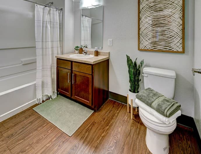 Modern bathroom in Fort Collins with wood flooring, a vanity with sink and mirror, toilet topped with towels and a potted plant, green rug, shower with white curtain, and framed artwork on the wall.