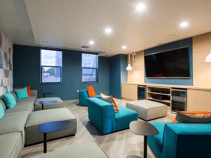 A modern lounge room in Fort Collins with blue and gray walls, teal and gray couches topped with orange and teal pillows, small black tables, a large flat-screen TV, and two windows letting in natural light.
