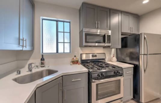 Modern kitchen with gray cabinets, stainless steel appliances including a gas stove, microwave, and refrigerator. A small window is above the sink, and a decorative bottle from the Shrine Collection adds charm to the white countertop.