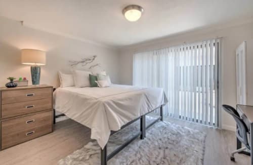 A modern bedroom with a raised bed, white bedding, a fur rug, and wooden dresser featuring Shrine Collection decor. Wall art and large sliding glass doors with vertical blinds fill the serene space with natural light.