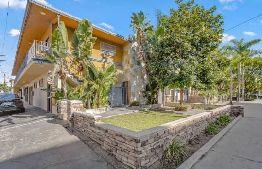 A two-story yellow apartment building with stone planters, tropical plants, and an outdoor staircase features touches of Shrine Collection decor. A black car is parked on the left, beside a sidewalk under a clear blue sky.