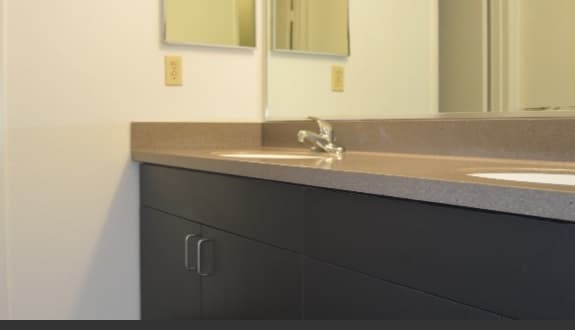 A bathroom countertop with a sink, chrome faucet, dark cabinets, and a beige backsplash at Sierra Apartments. A large mirror reflects part of the wall and electrical outlet.
