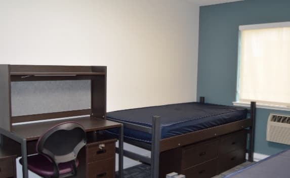 A dorm room in Sierra Apartments featuring a single bed frame and mattress, a desk with a chair, and drawers underneath the bed. There's a window with blinds, an air conditioning unit on the wall, and white and blue walls.
