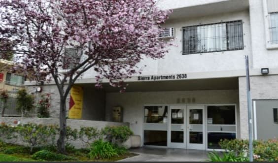 A beige rental apartment building with the sign Sierra Apartments 2638 above the entrance, glass doors, a flowering tree in front, and green shrubs along the walkway.