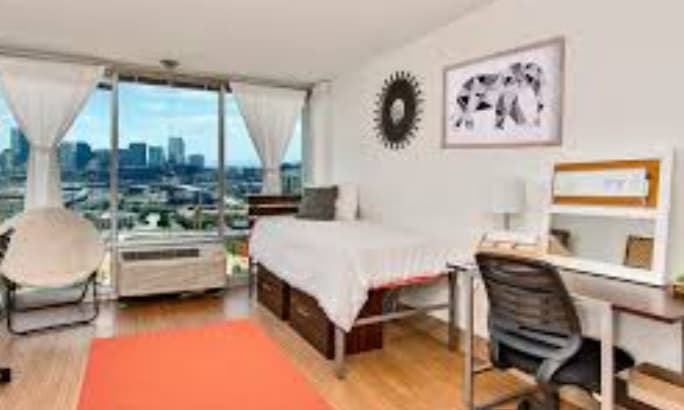 A bright, modern bedroom in Assembly Student Living with a single bed, desk, chair, and large windows offering a city view. Light wood floors, white walls, and contemporary decor create clean, airy student accommodation.