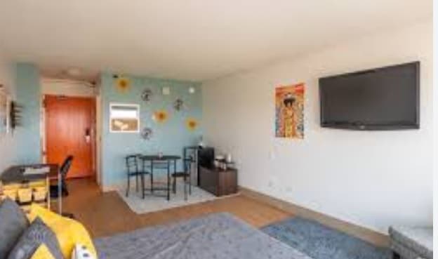 A bright studio apartment at Assembly Student Living with a wall-mounted TV, a small dining table for two, blue accent wall with floral decor, and wooden door. The room offers modern furnishings and natural light—ideal student accommodation.