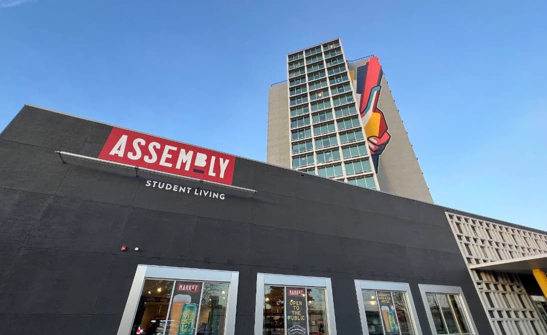 A modern building labeled Assembly Student Living offers vibrant student accommodation, with a tall tower behind it featuring a colorful mural of a hand holding a pencil set against the clear blue sky.