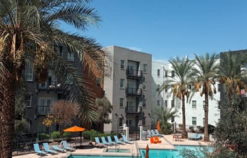 Sol Tempe Apartments in Tempe feature buildings with balconies overlooking a swimming pool area complete with lounge chairs, umbrellas, and palm trees under a clear blue sky.