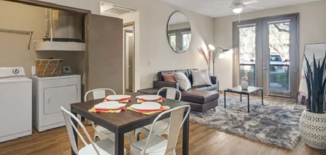 Modern Sol Tempe apartments with an open layout, featuring a dining table set for four, a cozy living area with sofa and rug, washer and dryer in a nook, large windows, and lush indoor plants.
