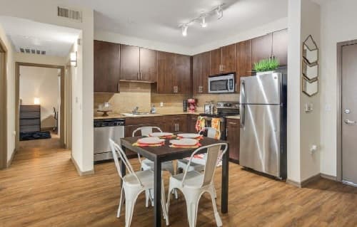 Modern kitchen at Sol Tempe Apartments with dark wood cabinets, stainless steel appliances, a small dining table for four with white chairs, wood flooring, and minimal decor. A hallway leads to another room in this Tempe home.