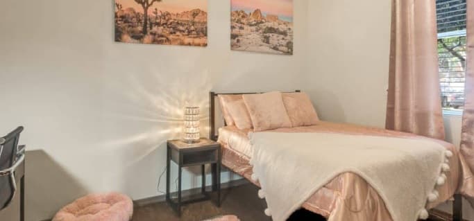 A cozy bedroom at Sol Tempe Apartments features a bed with pale pink bedding, a soft white throw, and pink pillows. A small nightstand with a lamp, pink furry rug, pink curtains, and desert landscape photos complete this Tempe retreat.