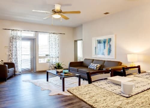 Bright, modern living room with a brown sofa, coffee table, armchair, and lamp. Large windows with sheer curtains fill this Saga Reno Reno space with light. Dining table with a mug and bowl in the foreground. Ceiling fan overhead.