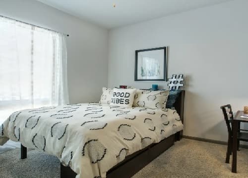 A neatly made bed with a white comforter featuring black circular patterns, a Good Vibes pillow, and framed art above it. Sunlight filters through sheer curtains after a recent Saga Reno Reno, with a desk and chair to the right.