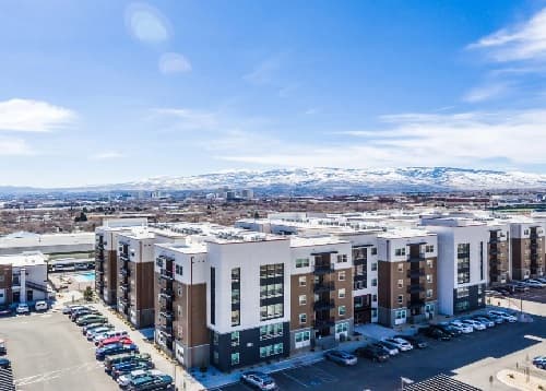 Aerial view of Saga Reno Reno’s modern apartment complex with parked cars in front, set against snow-capped mountains under a bright, blue sky.