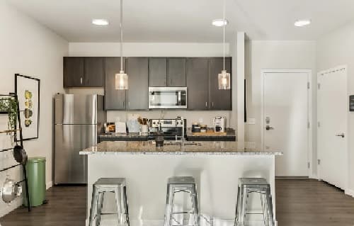 Modern kitchen at Stadium Apartments Fort Collins features dark cabinets, stainless steel appliances, a granite island with sink, three metal barstools, pendant lights, and minimalistic decor. White walls and wood flooring complete the space.