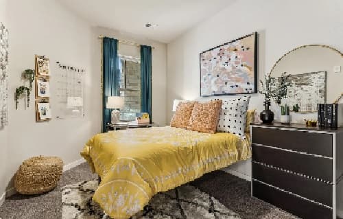 A cozy bedroom at Stadium Apartments Fort Collins features a yellow-patterned bedspread, pillows, a dark dresser with a round mirror, woven pouf, wall art, and a window with blue curtains letting in natural light.