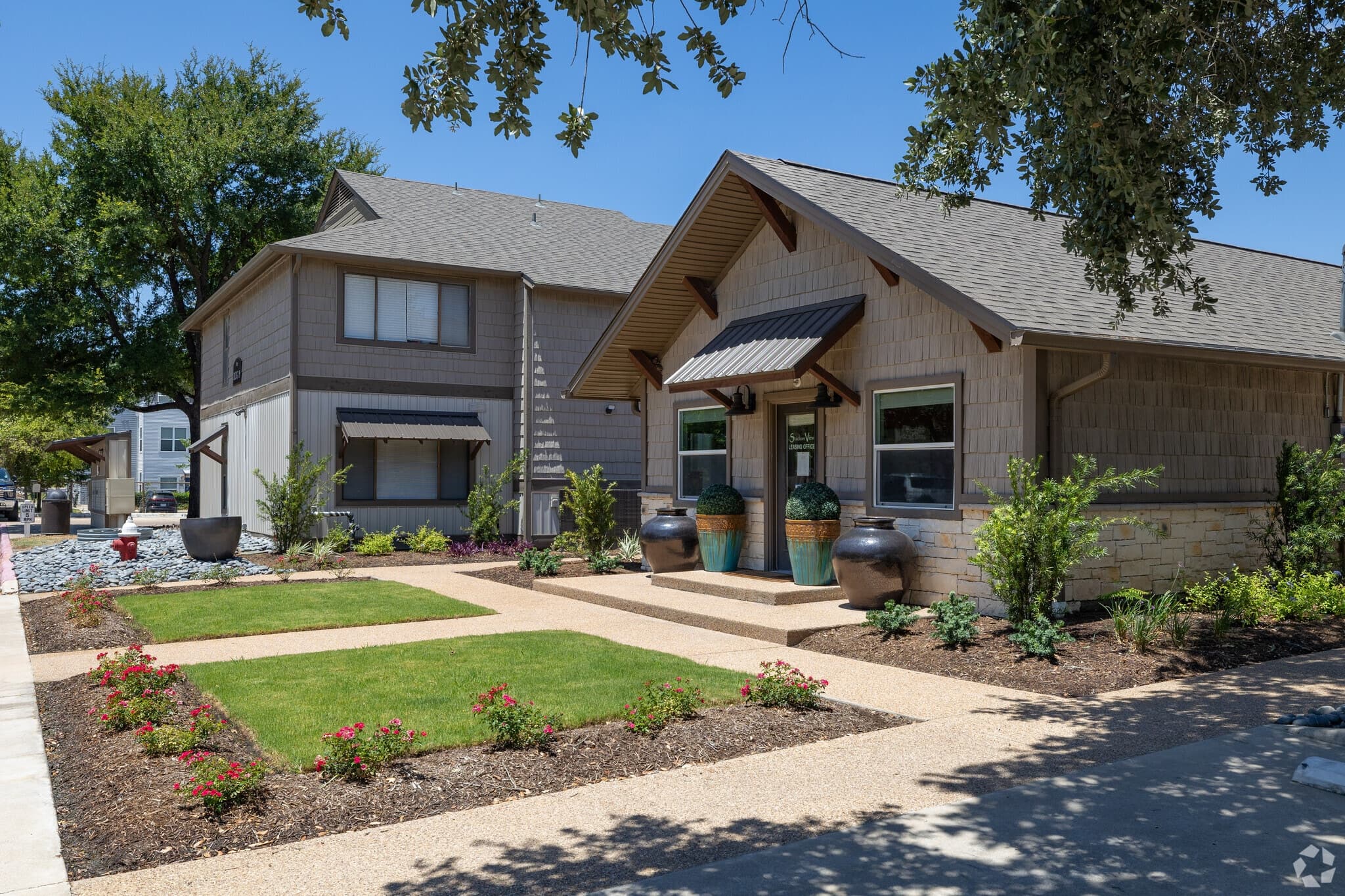 Modern suburban homes with manicured lawns and landscaped gardens, featuring large decorative pots near the entrance, offer a tranquil setting similar to Stadium View Apartments, all under a clear blue sky and surrounded by leafy trees.