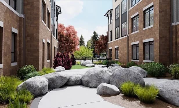 Landscaped courtyard at The Standard at Fort Collins, nestled between brick apartment buildings, features large rocks, green shrubs, trees with vibrant foliage, and a pathway leading to a fence and distant greenery under a partly cloudy sky.