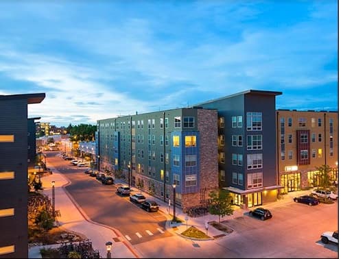 Modern apartment buildings line a quiet Fort Collins city street at dusk, with parked cars along the road, trees on the sidewalks, and warm lights glowing from windows and ground-floor businesses. The sky is partly cloudy.