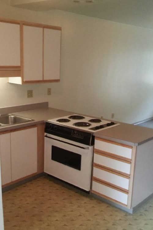 Small kitchen at Spencer View Apartments with light-colored cabinets, a white oven with four electric burners, stainless steel sink, and beige countertops. The floor features patterned linoleum and the walls are plain white.