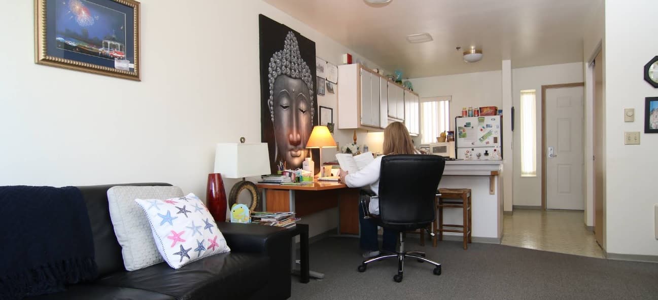 A woman sits at a desk in her cozy Spencer View Apartments home, reading papers. The room features a black couch, colorful pillows, large Buddha wall art, a kitchen area with a white fridge, and soft lighting.