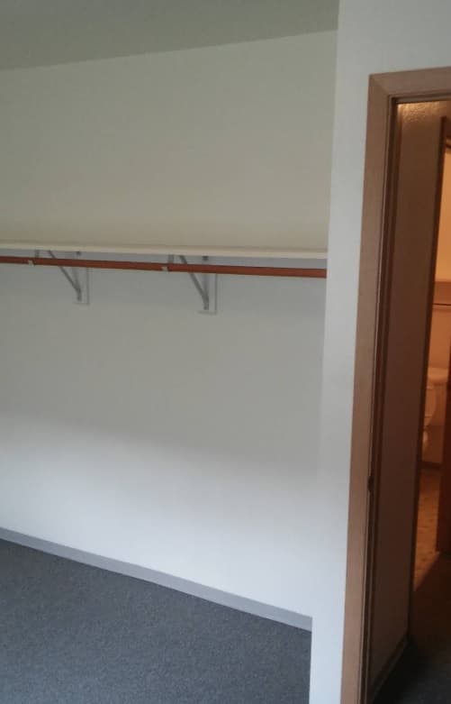 An empty closet with a single wooden shelf and hanging rod sits next to an open doorway leading to a bathroom at Spencer View Apartments. The floor is carpeted and the closet walls are plain white.