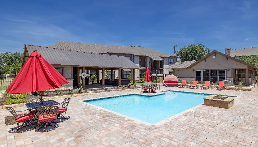 Outdoor pool area at Stadium View Apartments with lounge chairs, a small table and red umbrella, set among brick pavers and lush landscaping. Two-story buildings rise in the background below a clear blue sky.