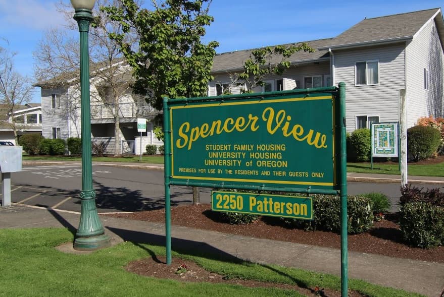 A green and yellow sign reads Spencer View Apartments, Student Family Housing, University Housing, University of Oregon and shows the address 2250 Patterson in front of white apartment buildings and a tree.
