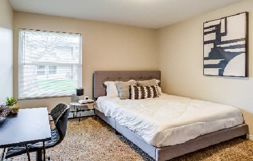Modern bedroom at The Social West® Fort Collins with a neatly made bed, patterned pillows, large window, abstract wall art, brown carpet, small desk with chair, and stylish decor. Natural light fills the room.