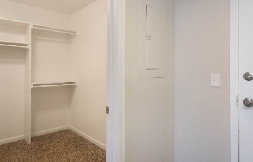Empty walk-in closet at The Social West® Fort Collins, featuring white shelves and hanging rods, beige carpet flooring, and a white wall with an electrical panel near the doorway.