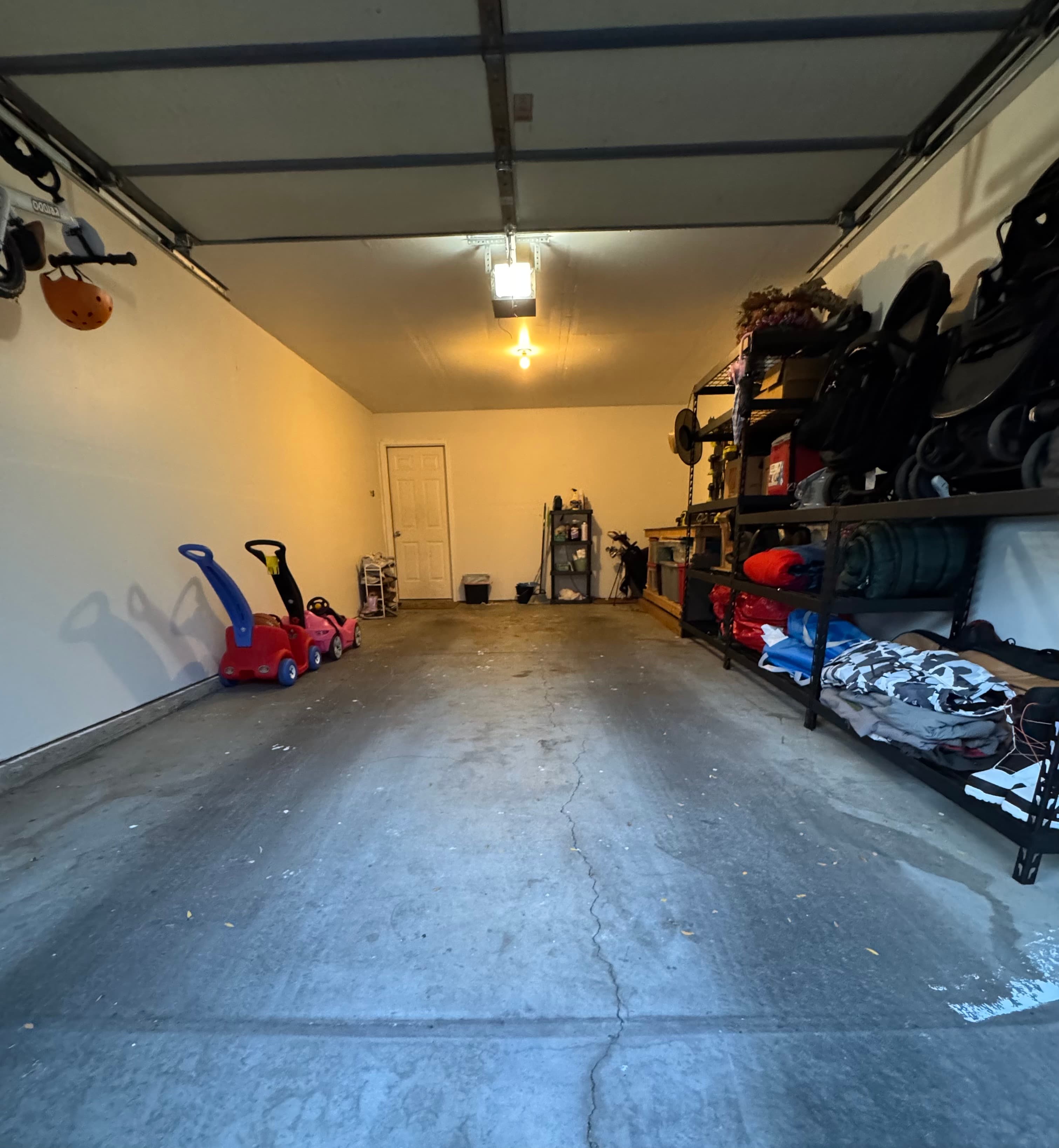 A clean, empty garage at 282 W 960 S, APT C features white walls, a concrete floor, and a closed overhead door. Shelves on the right store various items; two children’s ride-on toys rest against the left wall near a small table at the back.