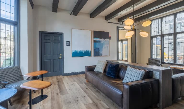 Modern lounge area with a dark leather sofa, wooden coffee tables, large windows, abstract art on the walls, and exposed ceiling beams. Natural light fills the space, creating a bright and inviting atmosphere.