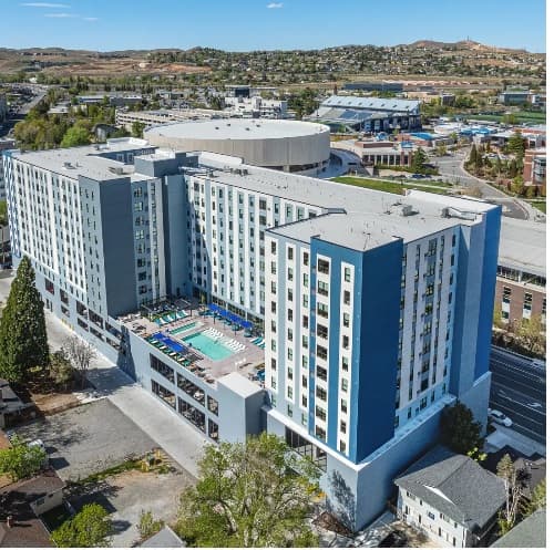 Aerial view of The Dean Reno, a modern multi-story apartment building with a central outdoor pool, surrounded by trees and roads, set in an urban area with hills in the background.