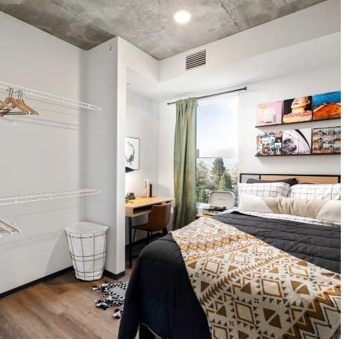 A modern, cozy bedroom at The Dean Reno features a bed with geometric bedding, a desk near a window with green curtains, wall shelves with books and decor, a laundry basket, and an open wire closet with hangers.