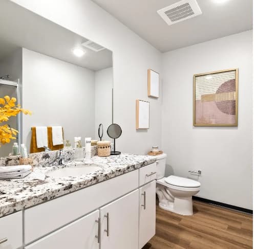 Modern bathroom at The Dean Reno with white cabinets, granite countertop, wood flooring, and wall art. Towels, soap, a mirror, and yellow flowers decorate the vanity. A toilet and holder are beside the counter.