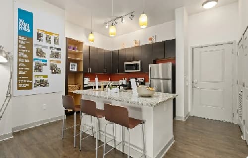 Modern kitchen at The District at Campus West Fort Collins with dark cabinets, stainless steel appliances, a granite island with three chairs, pendant lighting, wood flooring, white walls, and a stylish wall display featuring images and text.