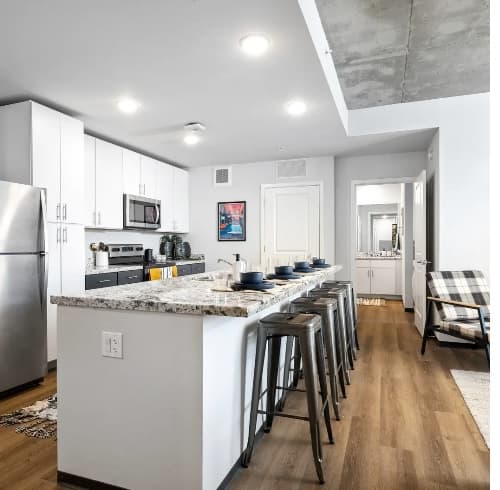 Modern kitchen at The Dean Reno features white cabinets, marble countertop island with five metal stools, stainless steel appliances, wood flooring, and a cozy seating area with a plaid chair. Bright lighting and a bathroom visible in the background.
