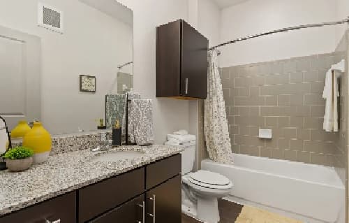 Modern bathroom at The District at Campus West Fort Collins featuring a granite countertop, dark wood cabinets, a sink with yellow vases, toilet, and bathtub with beige tiled wall. Towels and decorative items are neatly arranged for a stylish touch.