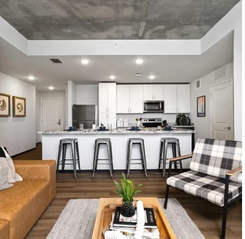 Modern open-concept kitchen and living area at The Dean Reno, featuring a white island, four metal barstools, stainless steel appliances, brown sofa, plaid accent chair, and a coffee table with decor on a gray rug.