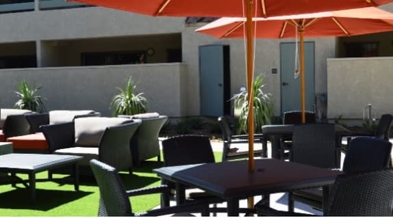 Outdoor patio area with dark wicker tables and chairs, shaded by large orange umbrellas, and surrounded by potted plants. The seating is set on artificial grass near a beige building with doors and balconies.
