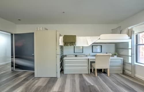 A modern bedroom at Telegraph Commons Apartments features a lofted bed above a white desk and drawers, a beige chair, framed artwork, and large windows letting in natural light. The space has light wood flooring and neutral-toned furniture.