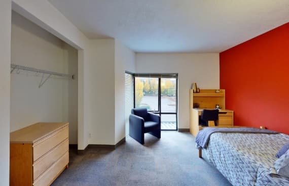 A clean, furnished dorm room with a bed, small desk, chair, dresser, open closet, and sliding glass door leading outside. One wall is painted red; the rest are white.