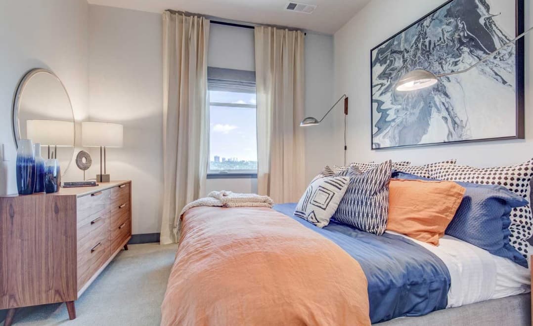 A modern bedroom with a bed featuring orange and blue bedding, decorative pillows, a wooden dresser with a round mirror, a window with curtains, and abstract wall art above the bed. A cityscape is visible outside the window.