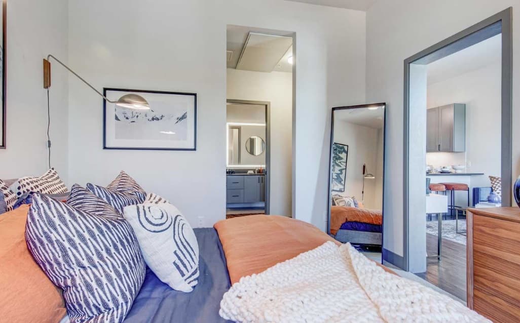A modern bedroom with a bed covered in patterned pillows and a rust-colored blanket, a wall lamp, framed art, a full-length mirror, and an open doorway leading to a bathroom and kitchen area.