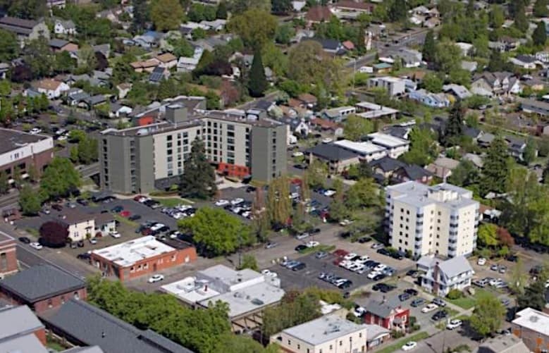 Aerial view of a suburban neighborhood with several mid-rise apartment buildings, smaller houses, parking lots, and tree-lined streets on a sunny day.