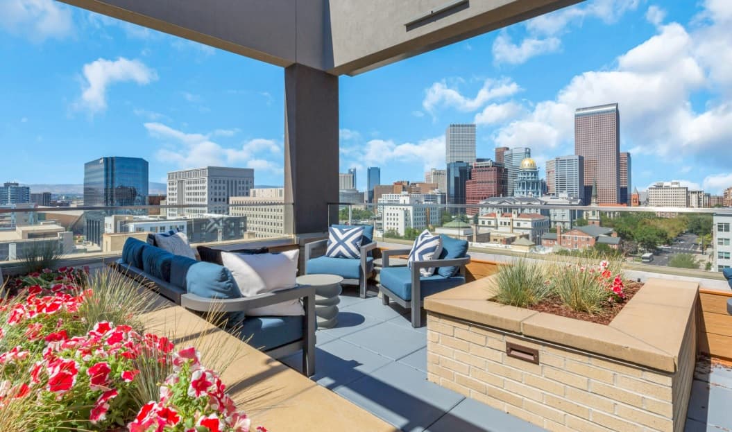 Rooftop patio with blue cushioned seating, small tables, planters with red and white flowers, and a city skyline with tall buildings under a blue sky with scattered clouds.